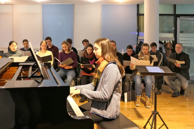 Studiochorprobe, im Vordergrund sitzt Studierende am Klavier, dahinter sitzt der Studiochor und singt