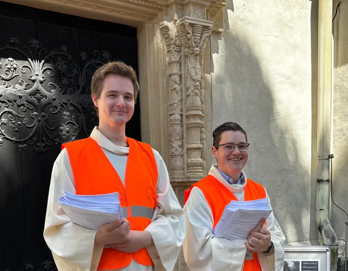 2 Studierende mit Warnwesten und Liedprogrammen am Eingang Dom